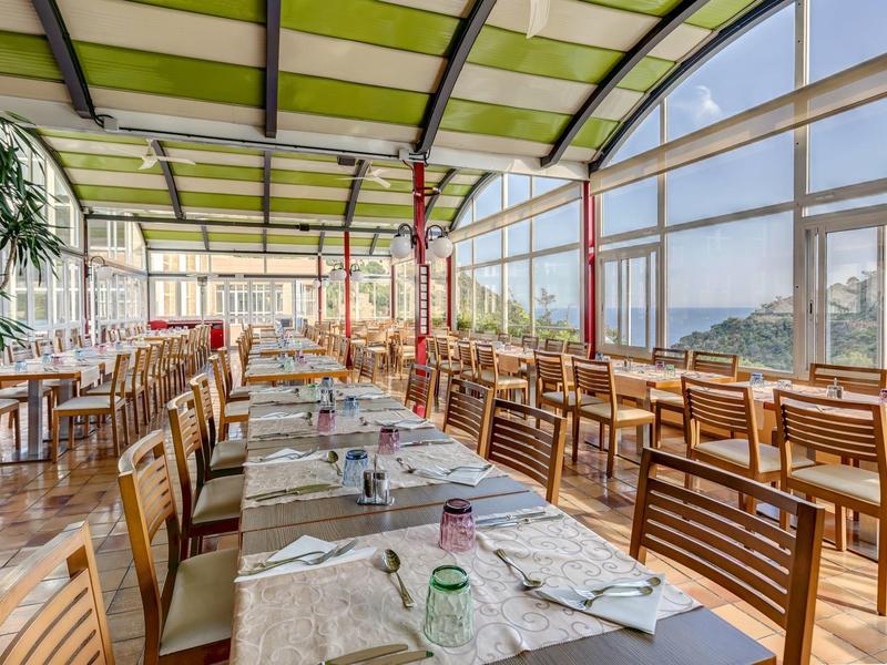 Bright hotel restaurant with large windows, wooden chairs, and green roofs overlooking mountains.