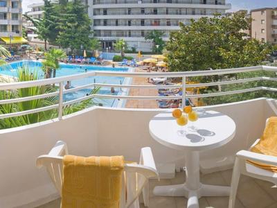 Balkon met twee stoelen, gele handdoeken en een tafel met drankjes met uitzicht op een hotelzwembad.