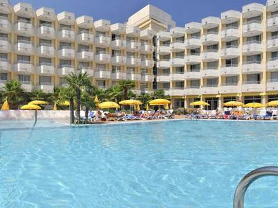 Hotelgebouw met veel balkons en een groot zwembad op de voorgrond bij zonnig weer.