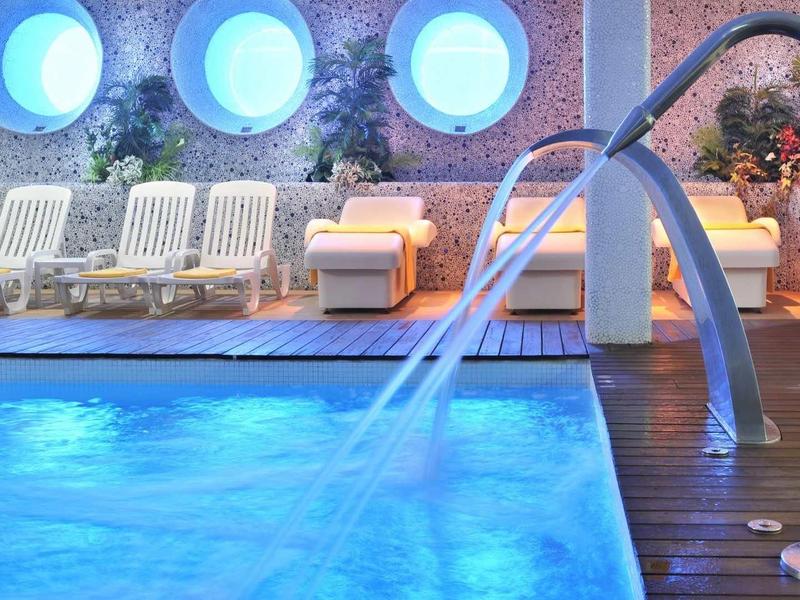 Binnenzwembad met blauwe verlichting, waterspelletjes en ligstoelen bij ronde ramen.