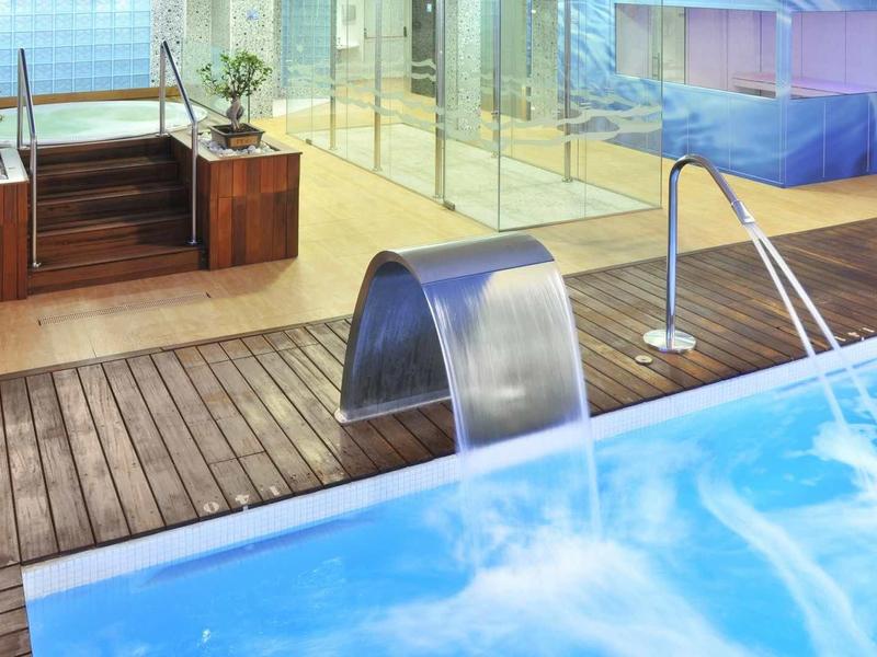 Moderne binnenzwembad met houten vloer, waterval douche en glazen afscheiding naar sauna.