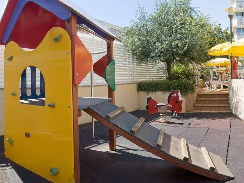 Speeltuin met klimrek en glijbaan naast hotelterras met parasols