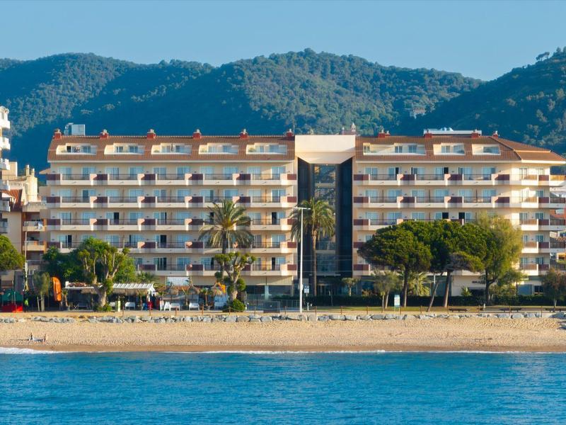 Edificio de hotel de varios pisos en la playa frente a colinas boscosas bajo cielo despejado.