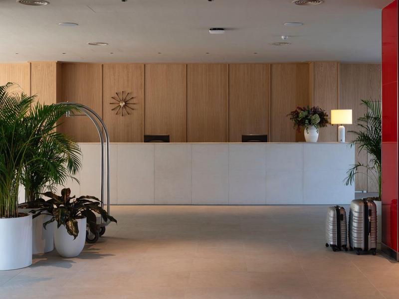 Recepción de hotel con mostrador blanco, plantas y ascensor rojo en el vestíbulo.