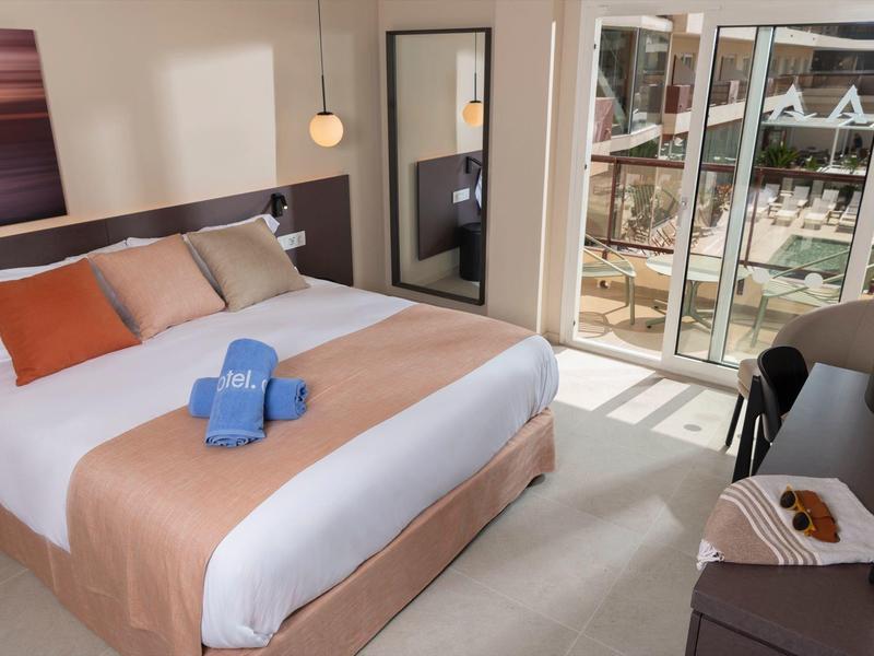 Habitación de hotel luminosa con cama grande, cojines coloridos y vista a un balcón soleado.