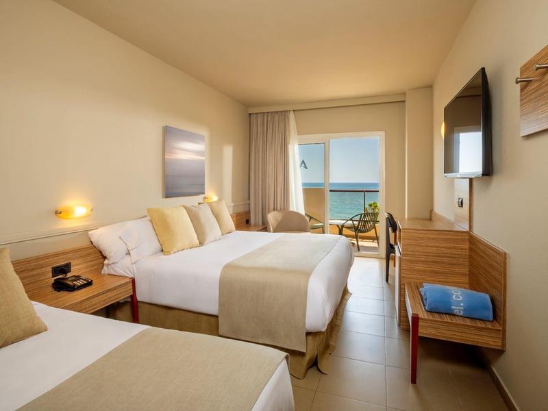 Habitación de hotel con dos camas dobles, balcón y vista al mar, moderna y luminosa.