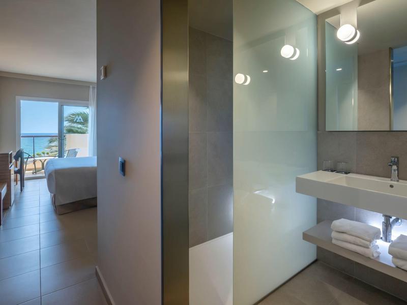 Área moderna de baño en hotel con ducha de vidrio, toallas y vista a la habitación luminosa.