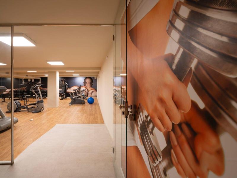 Área de fitness con máquinas y un espejo, personas entrenando en colchonetas y cintas de correr.