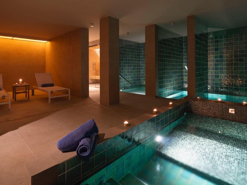 Área interior de hotel con jacuzzi iluminado y tumbonas relajantes con luz cálida.