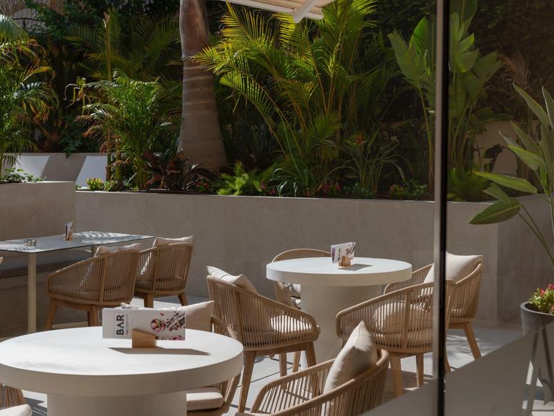 Área exterior moderna con mesas redondas y sillas rodeada de plantas en una esquina.