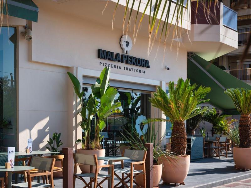 Entrada de un hotel con asientos, plantas grandes y sombra durante el día.