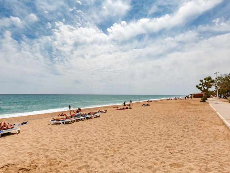 Amplia playa de arena con tumbonas y personas, mar azul y cielo nublado junto a un hotel.