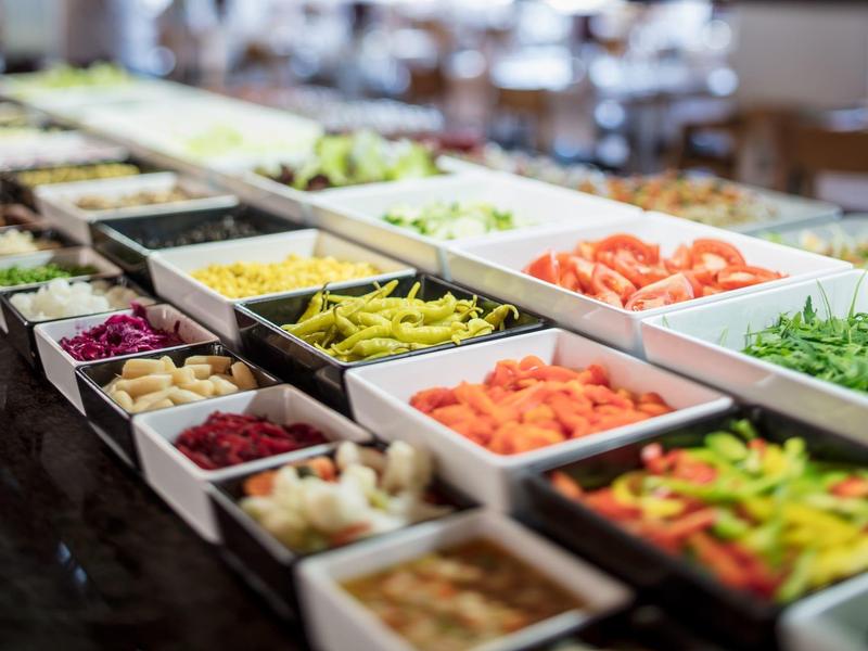 Buffet con diferentes verduras frescas en recipientes blancos sobre mesa oscura.