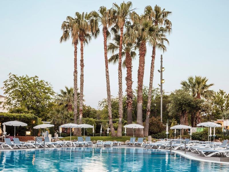 Großer Pool mit blauer Wasseroberfläche, umgeben von Liegestühlen, Palmen und Sonnenschirmen bei klarem Himmel.