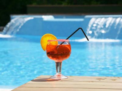 Ein rotes Cocktailglas mit Orangenscheibe und Strohhalm am Poolrand vor Wasserfall.