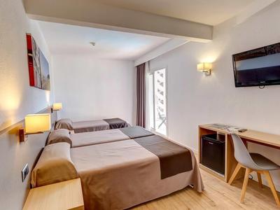 Hell eingerichtetes Hotelzimmer mit drei Betten, Schreibtisch und Wandfernseher.