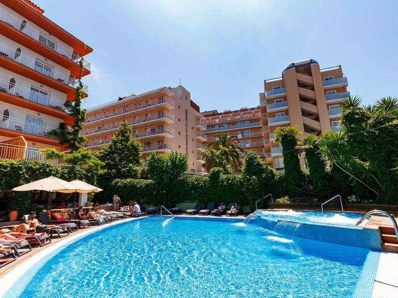 Outdoor pool in front of a multi-story hotel building on a sunny day with umbrellas and lounge chairs.