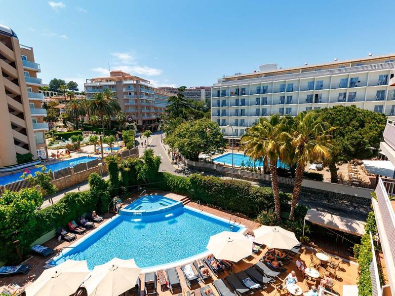 Hotel with outdoor pool, sun loungers, and palm trees under a sunny blue sky.