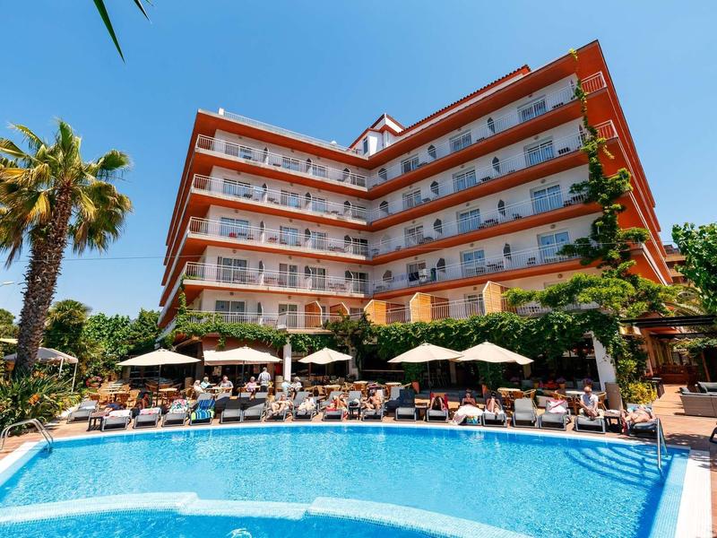 Multi-story hotel with pool and lounge chairs under blue sky and palm trees.