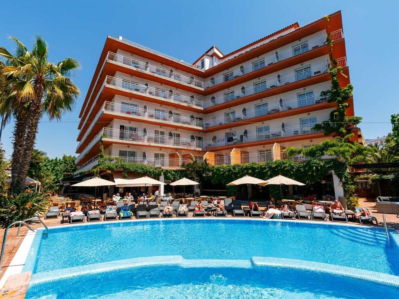 Hotel with large outdoor pool, sun loungers, and umbrellas surrounded by palm trees.