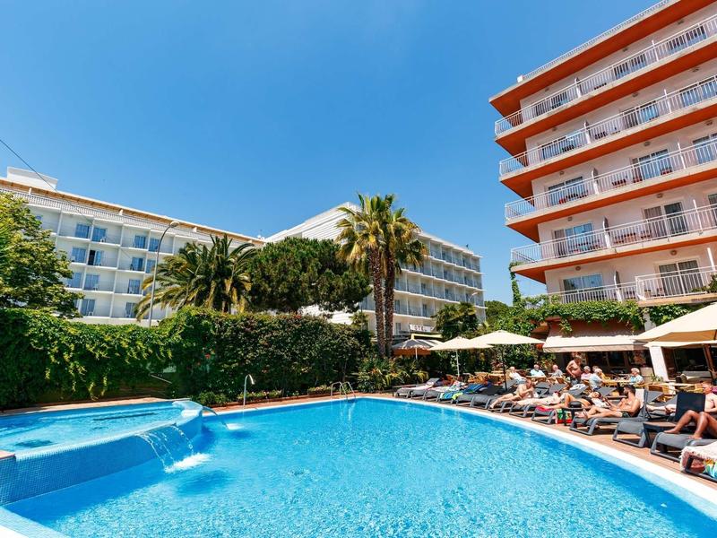 Hotel pool with sun loungers and umbrellas beside multi-story buildings under clear sky.
