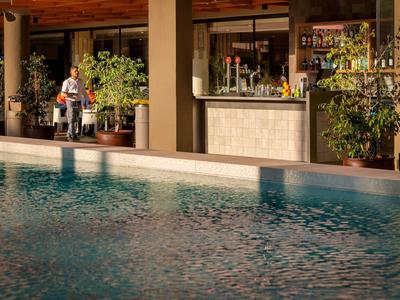 Hotelbar mit Sitzgelegenheiten neben einem blauen Swimmingpool, umgeben von Pflanzen.