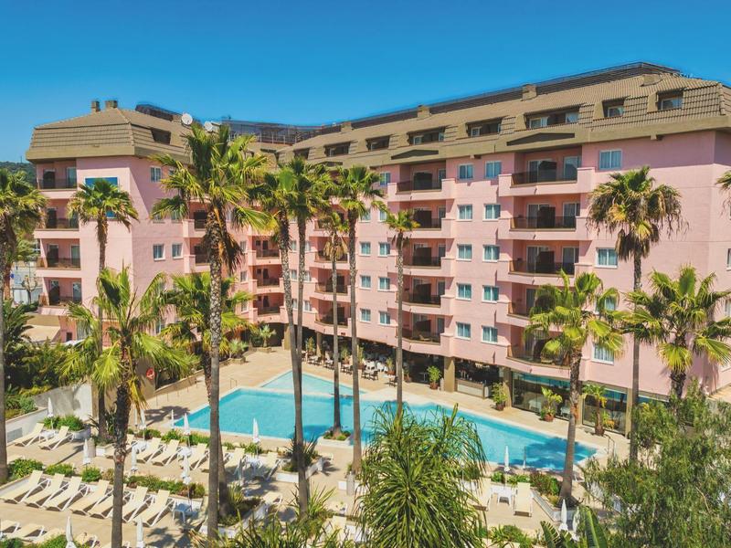Großes Hotel mit rosa Fassade, Palmen und einem langen Außenpool unter blauem Himmel.