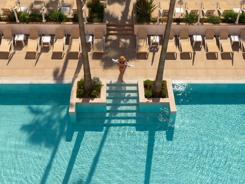 Großer blauer Pool mit Schatten von Palmen und beigen Liegestühlen auf heller Terrasse.