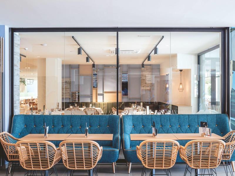 Modernes Café mit blauen Polstersofas, runden Holztischen und Korbstühlen vor großer Glasscheibe.