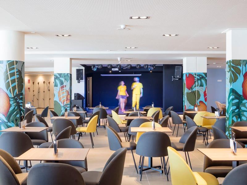 Moderner Veranstaltungsraum mit bunten Säulen, grauen Sesseln, gelben Sofas und Bühne im Hintergrund.