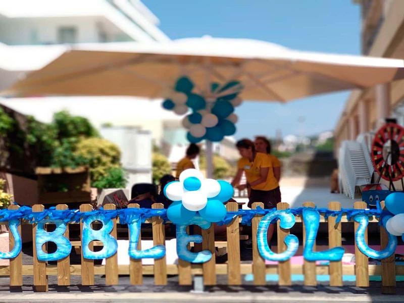 Bunte Außenwerbung mit blauer Schrift "BUBBLE CLUB" und blauen Luftballons an Holzzaun bei Sonnenschein.
