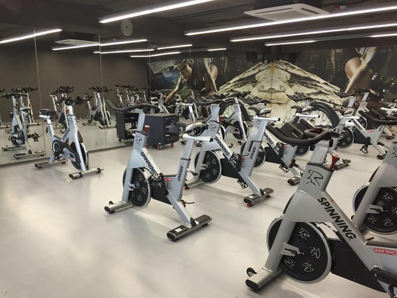 Fitnessraum mit mehreren grauen Spinning-Bikes, glattem Boden und Wandspiegeln im Hintergrund.