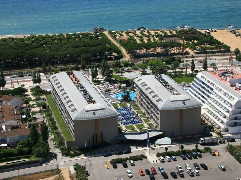 Zwei große Hotelgebäude mit Pool zwischen ihnen, Parkplätze und dahinter Strand und Meer sichtbar.