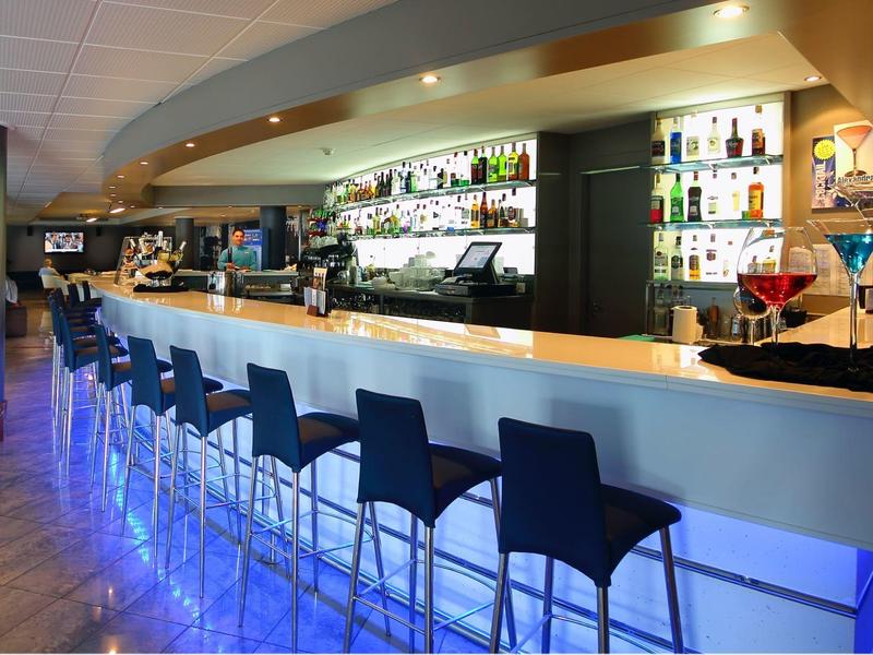 Moderne Bar mit blau beleuchtetem Tresen und schwarzen Barhockern in hellem Raum.