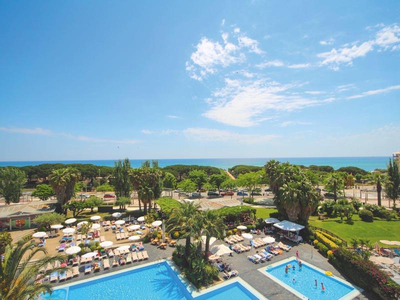 Blick auf große Poolanlage mit Sonnenschirmen, Palmen und blauem Himmel am Meer im Hintergrund.