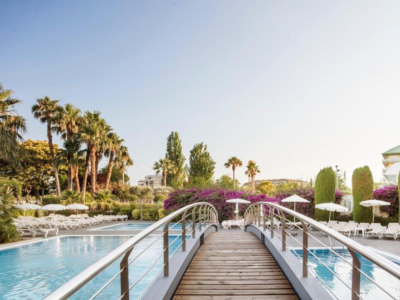 Holzbrücke über Pool mit Palmen, grünen Pflanzen und Liegestühlen bei klarem Himmel.