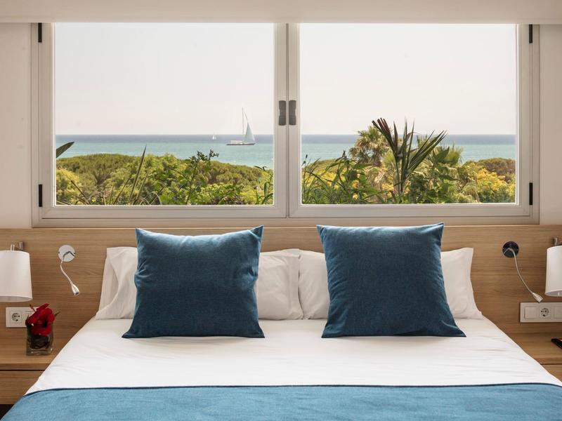 Schlafzimmer mit Doppelbett, blauen Kissen, seitlichen Lampen und Meerblick durch zwei große Fenster.