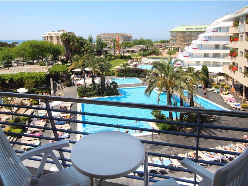 Balkon mit Tisch und Stühlen, Blick auf großen Pool, Palmen und Hotelgebäude bei klarem Himmel.
