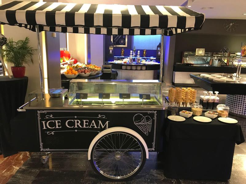 Eiswagen mit schwarz-weißer Markise und verschiedenen Eisbechern auf schwarzem Tisch daneben.