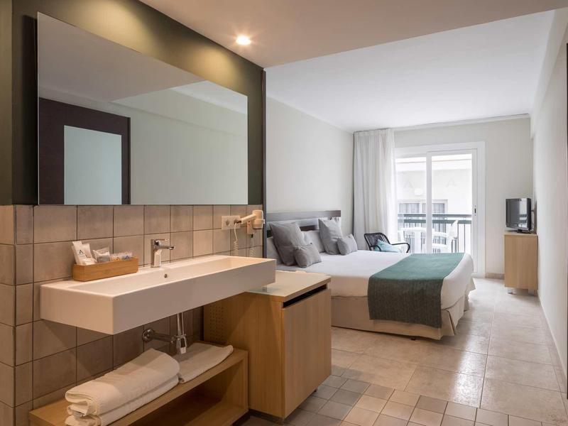Habitación de hotel luminosa con baño y cama doble, paredes grises claras y balcón