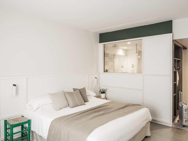 Habitación de hotel moderna con cama grande, colores neutros y taburete verde como acento.