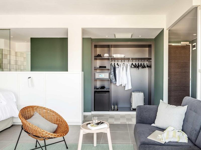 Habitación de hotel moderna con armario abierto, sofá gris y silla de mimbre en espacio luminoso.