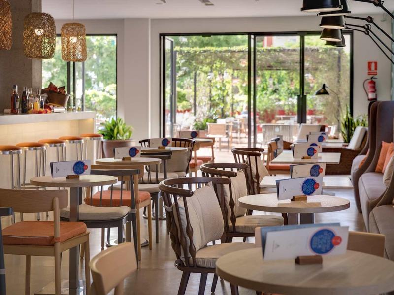 Cafetería luminosa con mesas redondas, sillas y grandes ventanas con vista al verde.