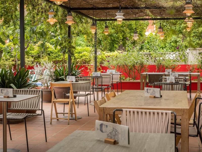 Área de terraza de hotel con mesas cubiertas, sillas y abundante vegetación verde.