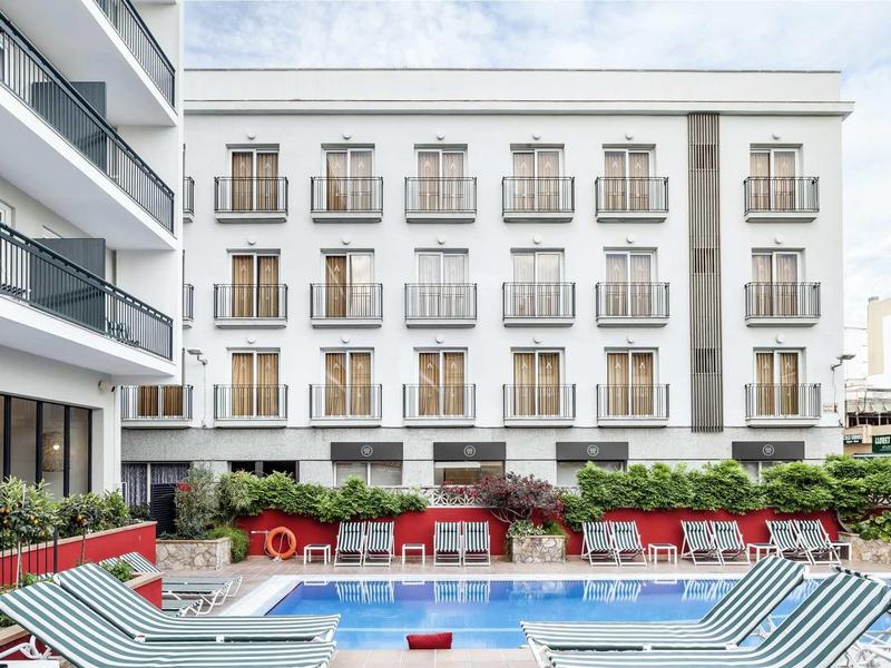 Hotel con piscina rodeada de tumbonas y un edificio blanco de varios pisos al fondo.