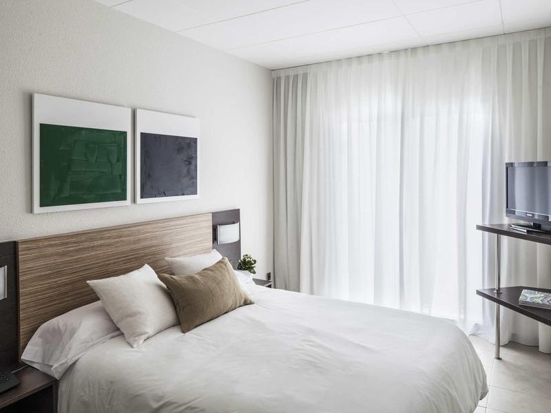 Habitación de hotel luminosa con cama doble, ropa de cama blanca, dos cuadros modernos y TV en la esquina.