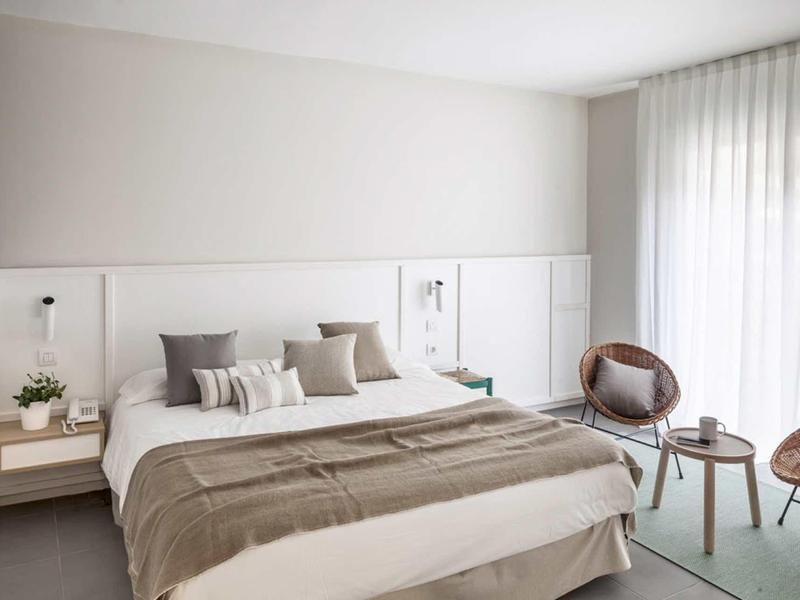 Habitación de hotel moderna con cama grande, mesitas, sillones y luz natural a través de cortinas.