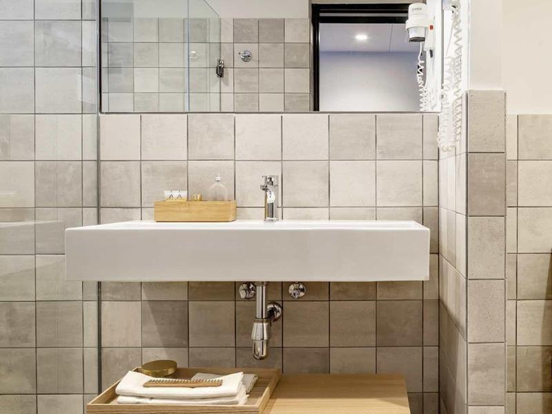 Baño moderno con azulejos grises cuadrados, lavabo blanco y mueble de madera con toallas.