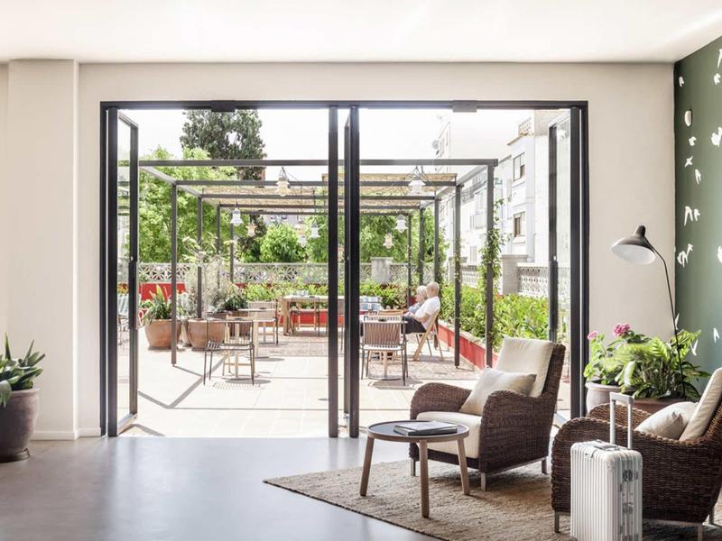 Zona de descanso luminosa en hotel con sillones cómodos, alfombra y vista a área exterior cuidada.