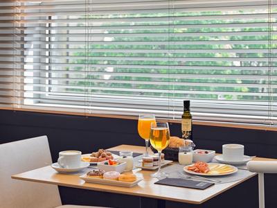 Kleiner Tisch mit Frühstücksgedeck, zwei Gläser Orangensaft, Weinflasche und Sitzplätze am Fenster mit Jalousien.